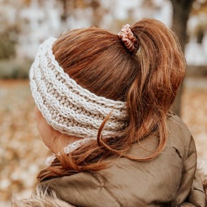 May include: A woman with long, red hair wearing a white knitted headband. Her hair is pulled back in a ponytail and secured with a pink scrunchie.