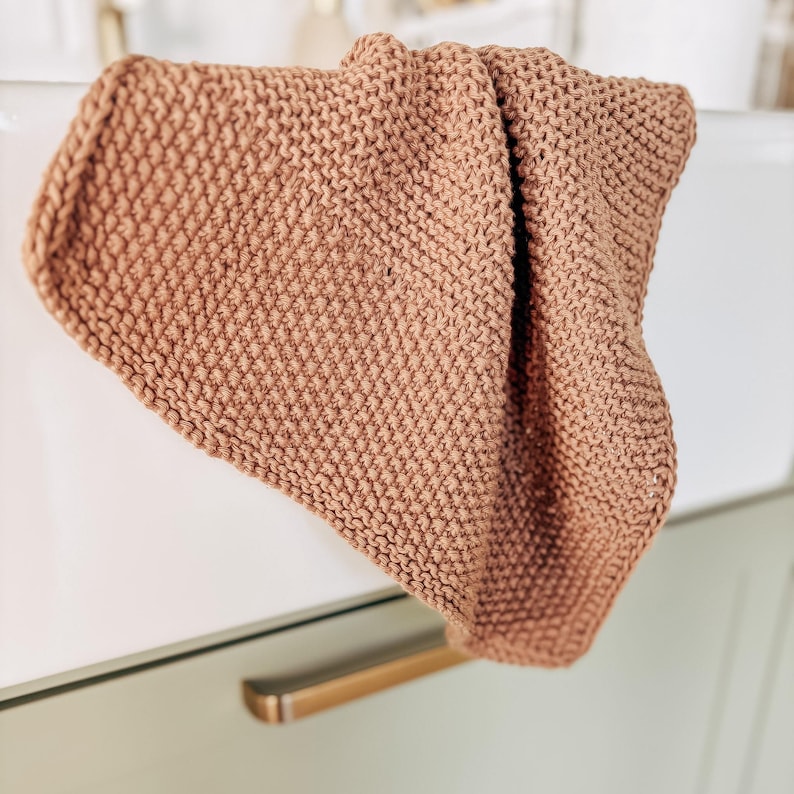 Op de afbeelding: Een vierkant, handgebreid washandje in een effen terracotta bruine kleur. Het textiel is voorzien van een consistent ribbelsteekpatroon, wat zorgt voor een getextureerd oppervlak met een afgewerkte, verstevigde rand langs de gehele omtrek.