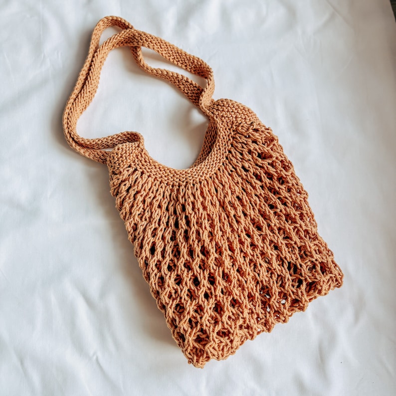 Peut inclure: Un sac fourre-tout en tricot, &agrave; tissage ouvert, de couleur marron chaud et terreux. Le sac est dot&eacute; d'une longue sangle int&eacute;gr&eacute;e pour &ecirc;tre port&eacute; &agrave; l'&eacute;paule. Le tissage ouvert cr&eacute;e un aspect en forme de filet, id&eacute;al pour transporter des courses ou d'autres articles.