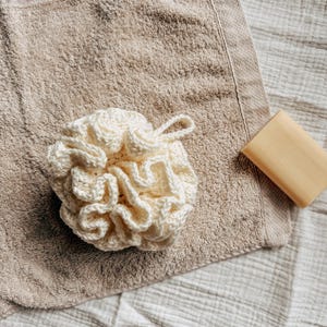 Può includere: Un pouf da bagno in crochet bianco su un asciugamano marrone. Il pouf ha la forma di un fiore con molti petali.