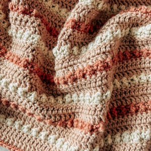 May include: A close-up of a crocheted blanket with a striped pattern in shades of pink, white, and orange.