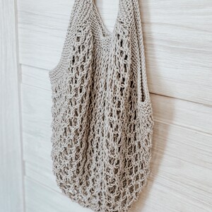 Peut inclure: Un sac fourre-tout beige tricot&eacute; avec un motif en forme de filet. Le sac a deux bretelles et une large ouverture. Le sac est suspendu sur un fond en bois de couleur claire.