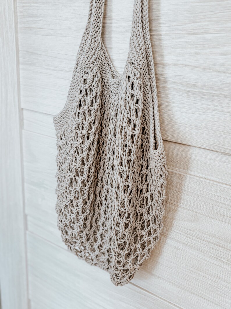 Peut inclure: Un sac fourre-tout beige tricot&eacute; avec un motif en forme de filet. Le sac a deux bretelles et une large ouverture. Le sac est suspendu sur un fond en bois de couleur claire.