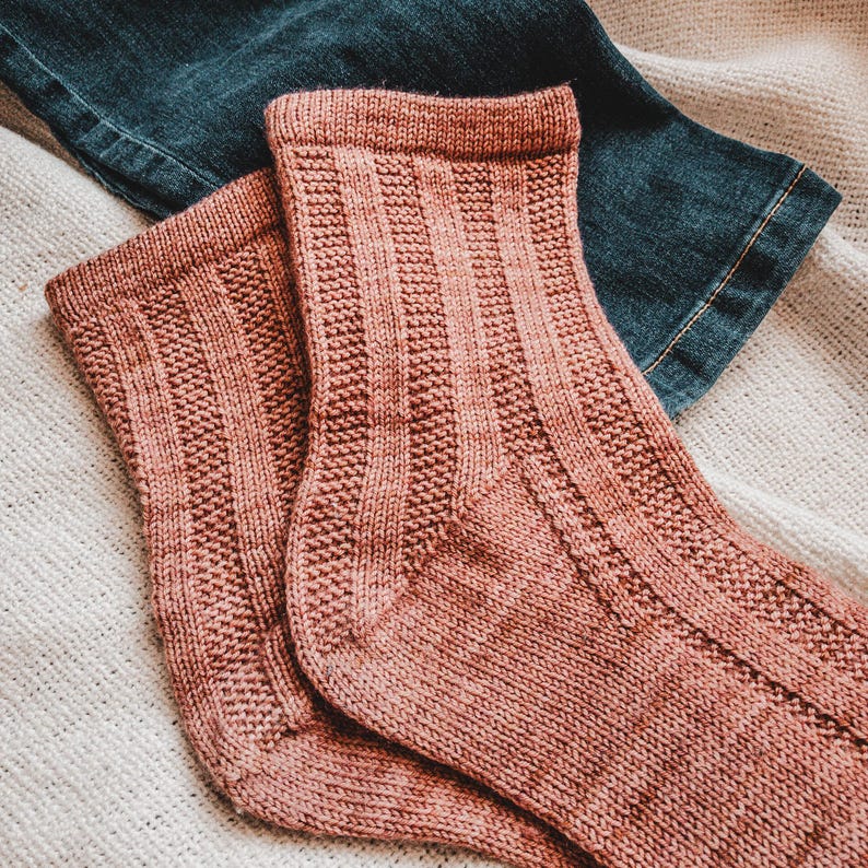 May include: A pair of knitted socks in a dusty rose color. The socks have a ribbed texture and are folded over at the top.