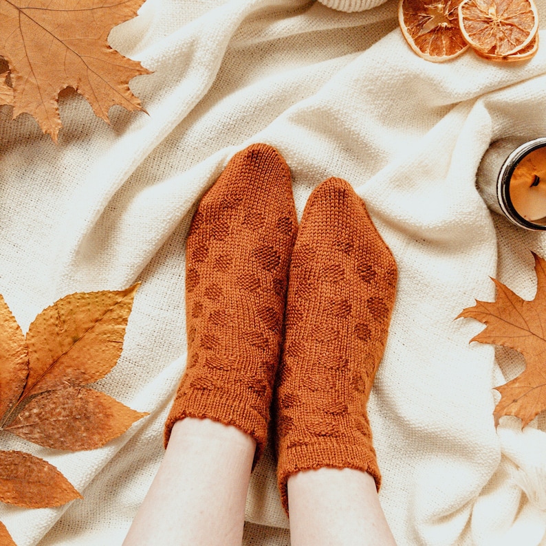 May include: A pair of rust-colored knitted socks with a geometric pattern. The socks are resting on a cream-colored blanket, surrounded by autumn leaves and dried orange slices. A lit candle is visible in the background.