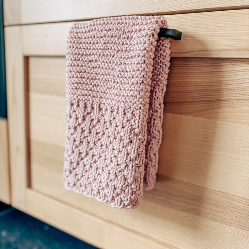 May include: A pink knitted dish towel hangs on a wooden cabinet door handle. The towel has a textured pattern and is folded in half.
