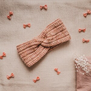 May include: A pink knitted headband with a twisted knot design. The headband is on a beige fabric background with pink bows scattered around it.