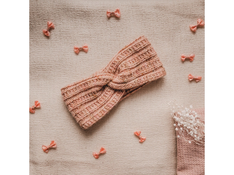 May include: A pink knitted headband with a twisted knot design. The headband is on a beige fabric background with pink bows scattered around it.