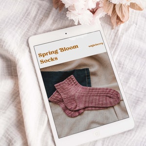 May include: A tablet displays a pair of pink knitted socks with the text "Spring Bloom Socks" and "whiljeberry". The socks are folded on a neutral-colored surface, with a denim fabric visible. Soft pink flowers are in the background.
