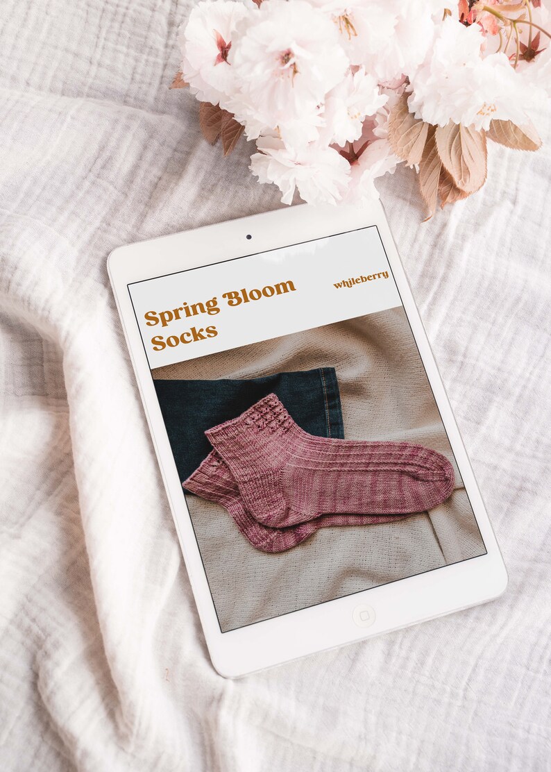 May include: A tablet displays a pair of pink knitted socks with the text "Spring Bloom Socks" and "whiljeberry". The socks are folded on a neutral-colored surface, with a denim fabric visible. Soft pink flowers are in the background.