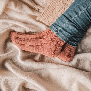 May include: A pair of knitted socks in a warm terracotta color. The socks are worn on a person's feet, with the toes of the socks visible. The socks are resting on a white blanket.