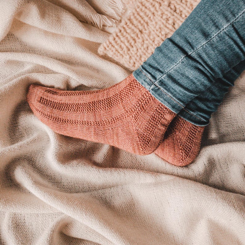 May include: A pair of knitted socks in a warm terracotta color. The socks are worn on a person's feet, with the toes of the socks visible. The socks are resting on a white blanket.