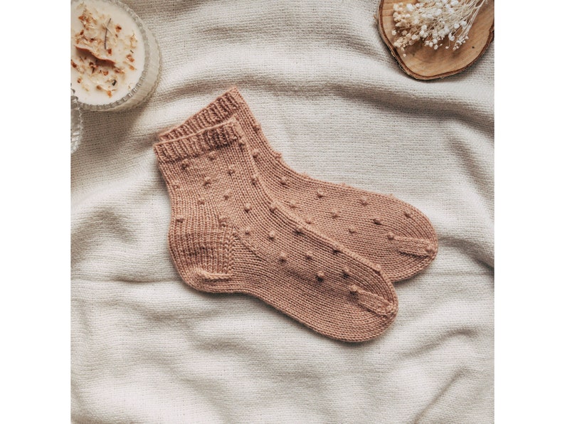 May include: A pair of knitted socks in a light brown color with a textured pattern of small dots. The socks are folded and laid out on a light beige fabric background.