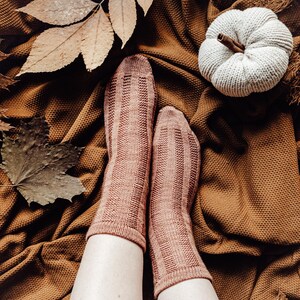 May include: A pair of brown knitted socks with a ribbed texture, worn on a person's feet. The socks are on a brown blanket with a white knitted pumpkin on top.