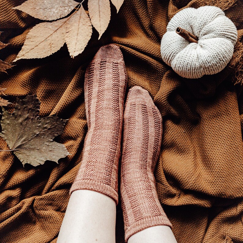 May include: A pair of brown knitted socks with a ribbed texture, worn on a person's feet. The socks are on a brown blanket with a white knitted pumpkin on top.