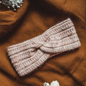 May include: A pink knitted headband with a twisted knot design. The headband is on a brown textured surface.