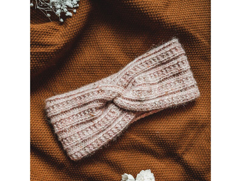 May include: A pink knitted headband with a twisted knot design. The headband is on a brown textured surface.