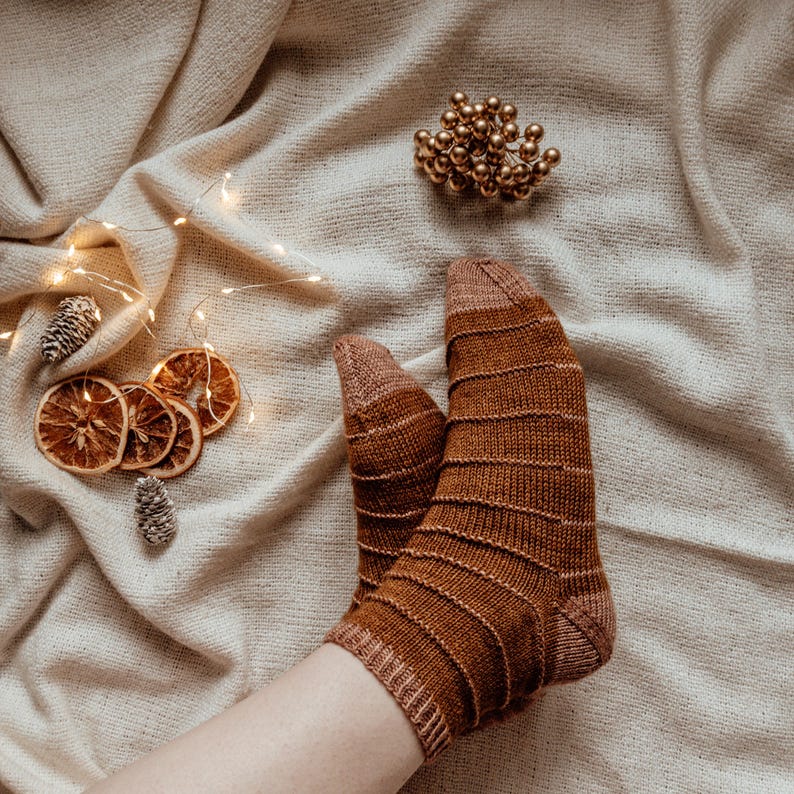 Puede incluir: Un par de calcetines de punto marr&oacute;n con un patr&oacute;n de canal&eacute;, descansando sobre una manta de color crema con luces de cadena y rodajas de naranja secas.