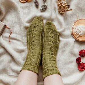 May include: A pair of green knitted socks with a cable knit pattern. The socks are on a white fabric background.