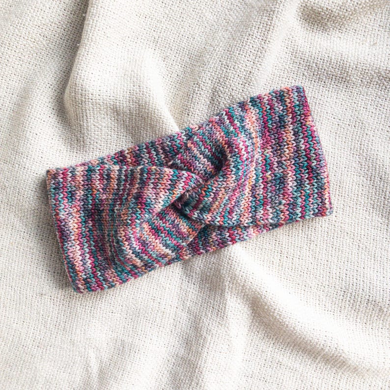 May include: A multi-colored knitted headband with a twisted knot design. The headband is made of a soft, stretchy yarn and features a variety of colors, including pink, purple, blue, and green.