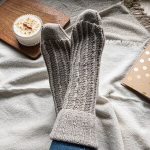 May include: A pair of light gray, ribbed knit socks. The socks are resting on a cream-colored blanket. A small wooden cutting board with a candle and a book with gold polka dots are also visible.