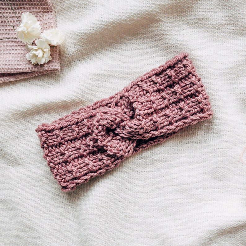 May include: A pink knitted headband with a knotted design. The headband is made of a soft, textured yarn.