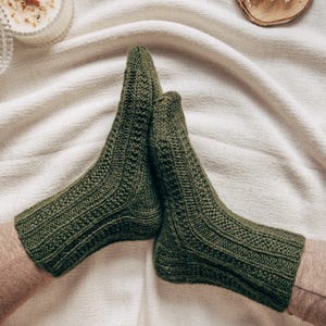 Op de afbeelding: Een paar groene gebreide sokken met een kabelbreipatroon. De sokken zijn dubbelgevouwen en liggen op een wit stofoppervlak.
