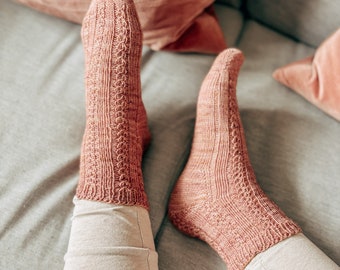 Peut inclure: Une paire de chaussettes en tricot rose avec un motif en câble. Les chaussettes sont portées aux pieds d'une personne et reposent sur un canapé gris.