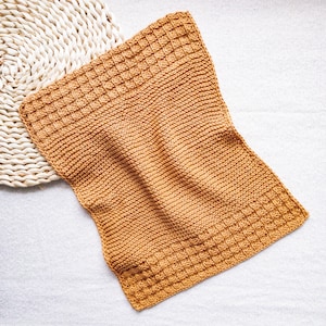 May include: A golden-brown knitted washcloth with a textured, waffle-like pattern at the top and bottom edges. The washcloth is rectangular and lies on a white surface next to a woven, circular placemat.