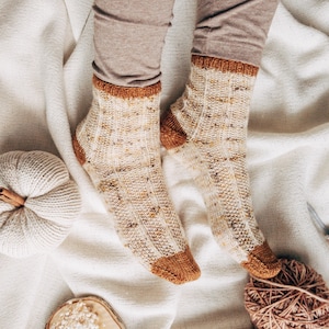 May include: A pair of beige and brown knitted socks with a speckled pattern. The socks are on a white blanket with a white pumpkin and a ball of yarn.
