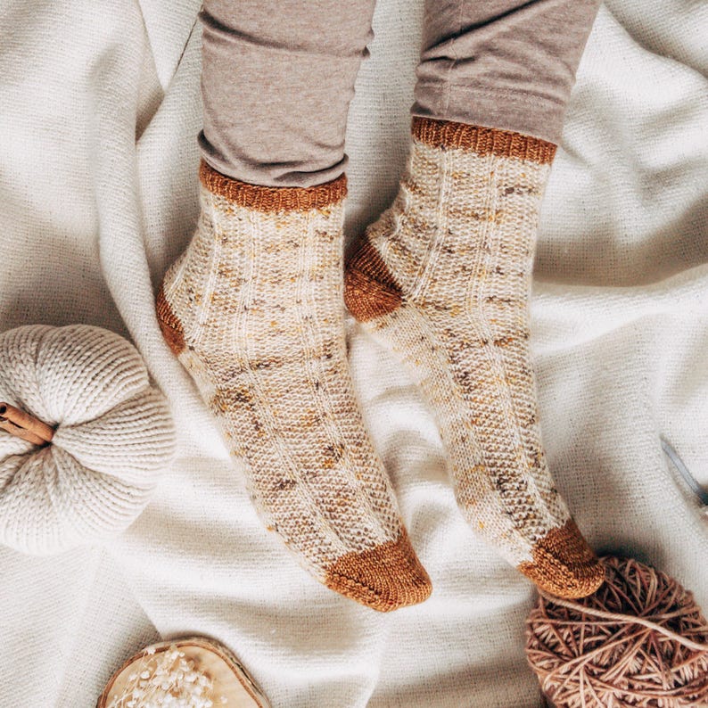 May include: A pair of beige and brown knitted socks with a speckled pattern. The socks are on a white blanket with a white pumpkin and a ball of yarn.