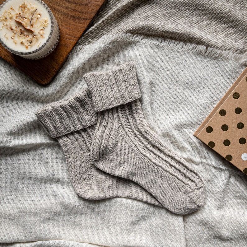 May include: A pair of light gray knitted socks with ribbed detailing, folded over at the top. The socks are lying on a textured, off-white blanket. A candle in a glass jar and a small gift box with gold polka dots are also visible.