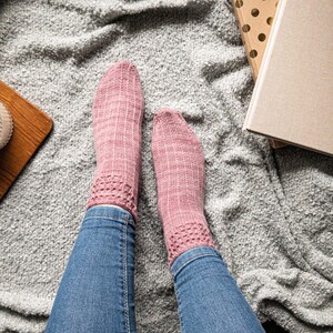 May include: A pair of pink knitted socks with a decorative openwork pattern at the ankle. The socks are worn with blue jeans, resting on a gray textured blanket. A wooden tray and books are in the background.