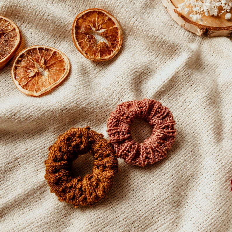 K&ouml;nnte beinhalten: Zwei geh&auml;kelte Haargummis, eines braun und eines rosa, auf einem wei&szlig;en Stoffhintergrund. Getrocknete Orangenscheiben sind ebenfalls sichtbar.