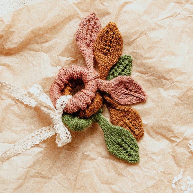 May include: A set of four knitted hair scrunchies in shades of pink, green, and brown. The scrunchies are arranged in a flower shape on a brown paper background.