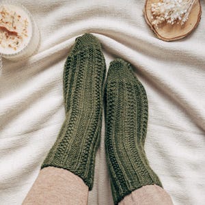May include: A pair of green knitted socks with a ribbed texture. The socks are worn on a person's feet and are resting on a white blanket.