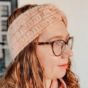 May include: A pink knitted headband with a twisted design. The headband is worn by a person with brown hair and glasses.