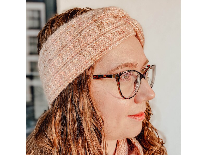 May include: A pink knitted headband with a twisted design. The headband is worn by a person with brown hair and glasses.