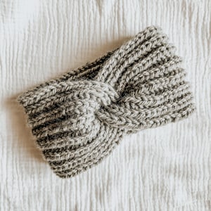 May include: A grey knitted headband with a knotted design. The headband is made from a soft, textured yarn.