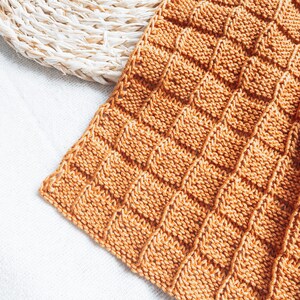 May include: A close-up of a knitted throw blanket in a warm orange color. The blanket has a textured, waffle-like pattern.