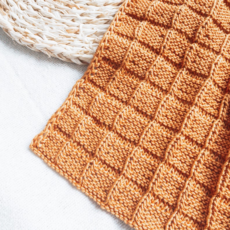 May include: A close-up of a knitted throw blanket in a warm orange color. The blanket has a textured, waffle-like pattern.