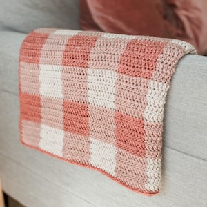 May include: A pink and white crocheted throw blanket with a gingham pattern. The blanket is draped over a gray couch.