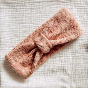 May include: A pink and white knitted headband with a knotted design. The headband is made of a soft, fuzzy yarn.