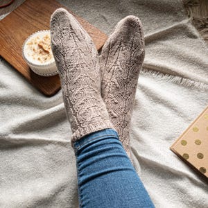 May include: A pair of beige knitted socks with a detailed cable pattern. The socks are worn with blue jeans. A wooden cutting board with a small bowl of food and a small gift box are in the background.