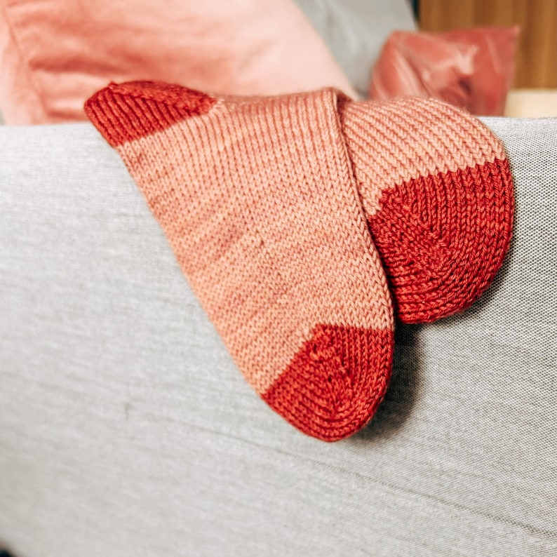 K&ouml;nnte beinhalten: Ein Paar gestrickte Socken mit einem rosa-roten Streifenmuster. Die Socken sind gefaltet und liegen auf einer grauen Stoffoberfl&auml;che.