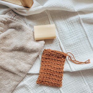 May include: A brown knitted soap sack with a drawstring closure, a bar of soap, and a beige towel on a white textured surface.
