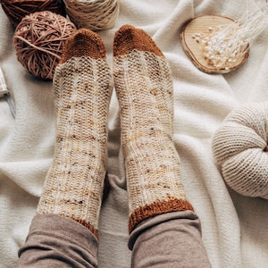May include: A pair of knitted socks with a brown and beige color scheme. The socks have a ribbed pattern and are worn on a person's feet. The socks are on a white blanket with a knitted pumpkin and yarn balls.