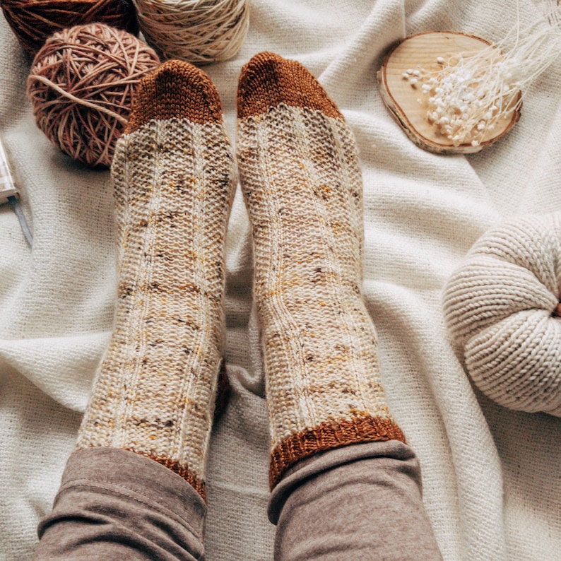 May include: A pair of knitted socks with a brown and beige color scheme. The socks have a ribbed pattern and are worn on a person's feet. The socks are on a white blanket with a knitted pumpkin and yarn balls.