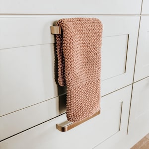 Op de afbeelding: Een oudroze handgebreide handdoek met een textuur van ribbelsteek, gedrapeerd over een handdoekhouder van geborsteld metaal. De handdoek is rechthoekig van vorm en heeft een consistente breistructuur over het gehele oppervlak van de stof.