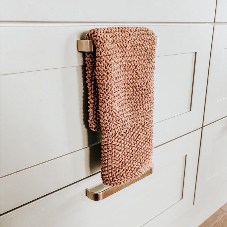 Op de afbeelding: Een oudroze handgebreide handdoek met een textuur van ribbelsteek, gedrapeerd over een handdoekhouder van geborsteld metaal. De handdoek is rechthoekig van vorm en heeft een consistente breistructuur over het gehele oppervlak van de stof.